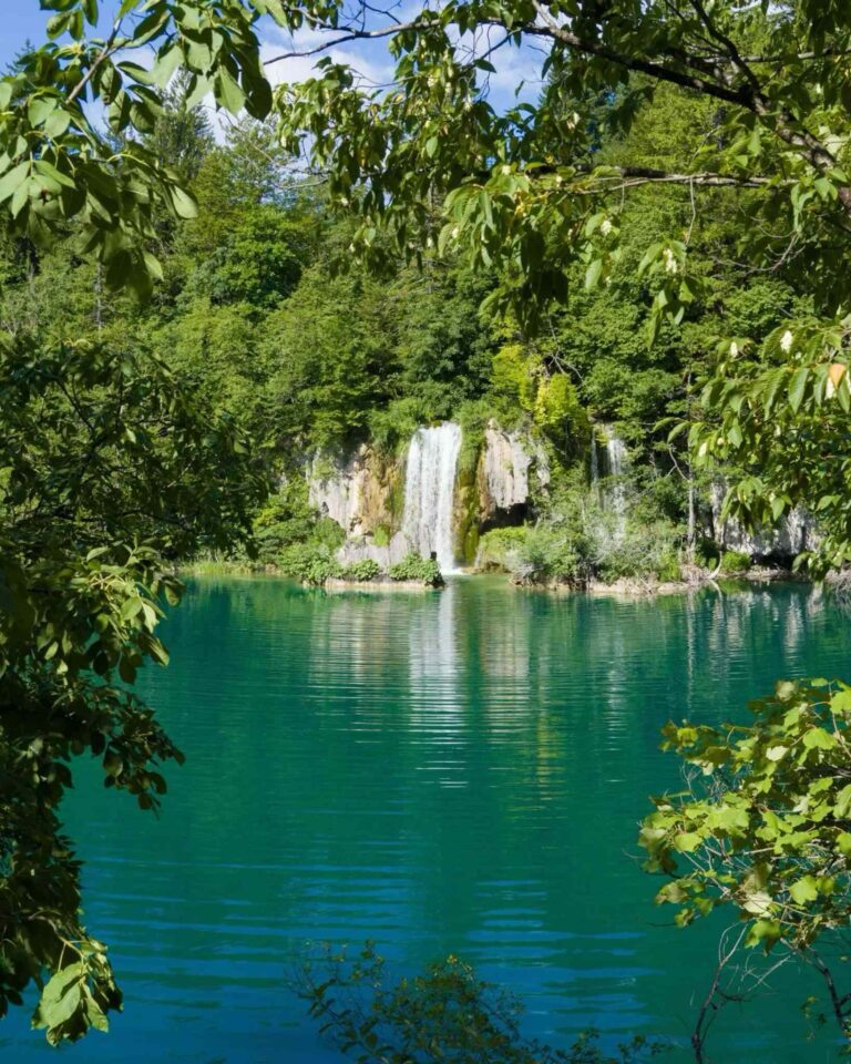 Waterfall flowing into a turquoise lake at Plitvice Lakes National Park, framed by green forest.