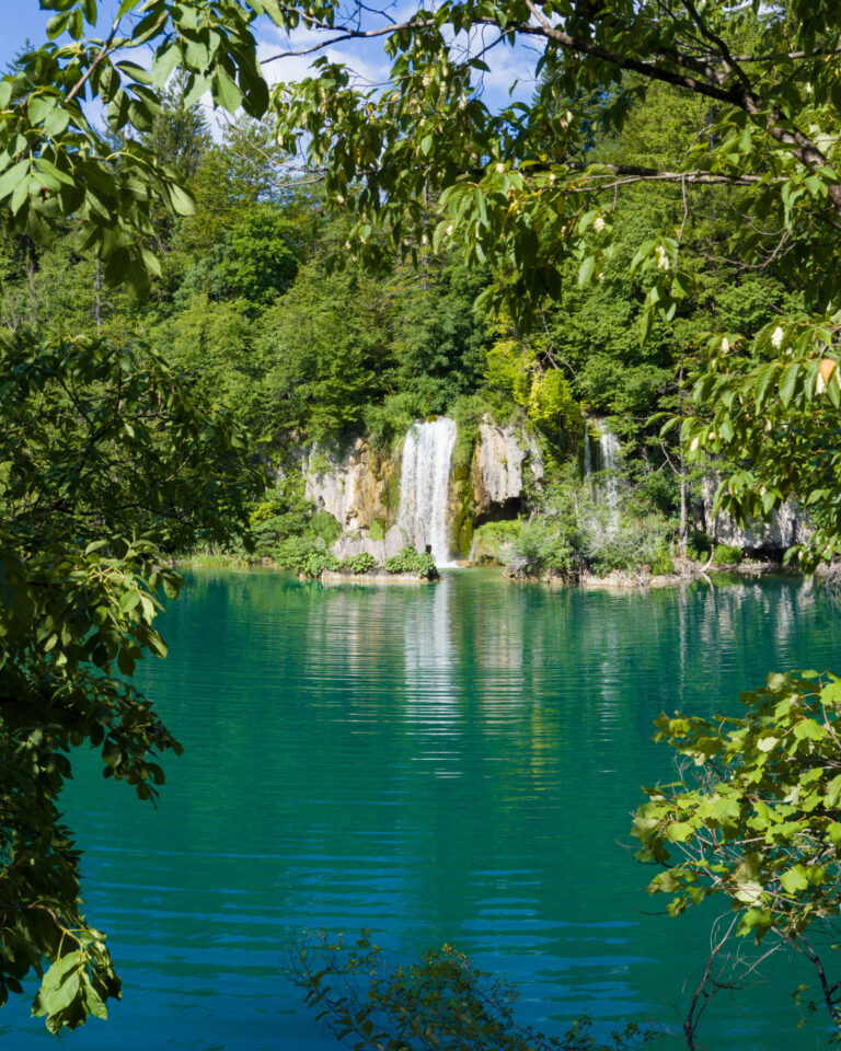 Waterfall flowing into a turquoise lake at Plitvice Lakes National Park, framed by green forest.